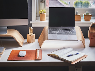 Home office with monitor and laptop resting on light wood stands. Notebook, pen, mouse, and mousemat also on top of white surface desk. 3 small plants visible behind on windowsill. 