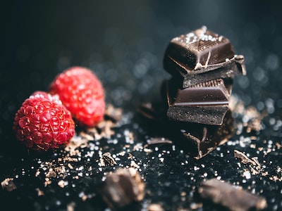 two raspberries and three squares of dark chocolate on a black surface, surrounded by crumbs of sugar/salt, and chocolate shavings