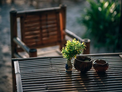 An out of focus wooden garden chair, behind a bamboo table with two bowls and a green plant on top. The plant is in a small white vase. In the background there is also an out of focus green plant.