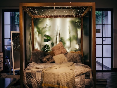 Wooden bedframe with fairy lights and plants behind screen. Tall mirror to left of bed. 