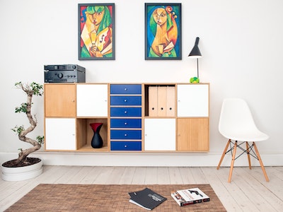 Picture of room with colourful artwork, cabinet with blue drawers, chair to the right and books on the floor.